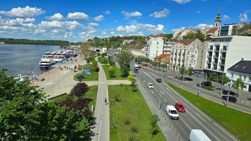 View from Branko's Bridge in Belgrade, overlooking the Sava River waterfront with docked international ships, the lively Beton Hala promenade, and the Kalemegdan Fortress and Saint Michael's Cathedral rising in the background.